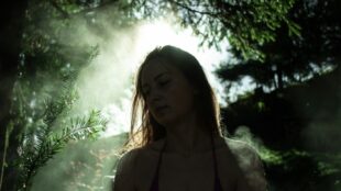 Kvinna i skogen omgiven av solljus och dis, med antydan till sång