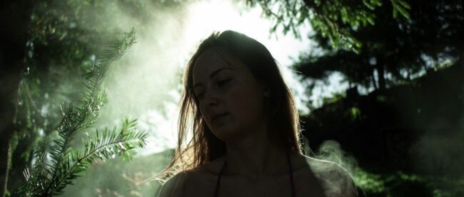 Kvinna i skogen omgiven av solljus och dis, med antydan till sång